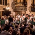 A choir performs in a historic church, led by a conductor, with a seated audience applauding enthusiastically.