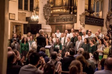 A choir performs in a historic church, led by a conductor, with a seated audience applauding enthusiastically.