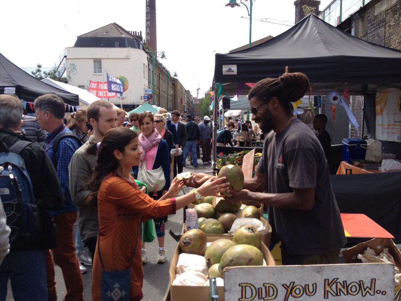 A bustling scene at Brick Lane market, with a vendor selling coconuts to a customer, surrounded by vibrant market stalls.