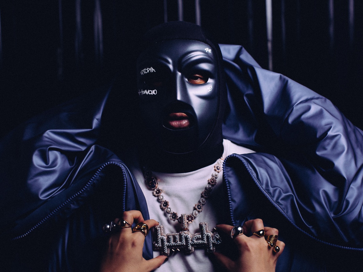 M Huncho in his signature mask and bold jewelry, showcasing his unique style and enigmatic persona in this striking portrait.