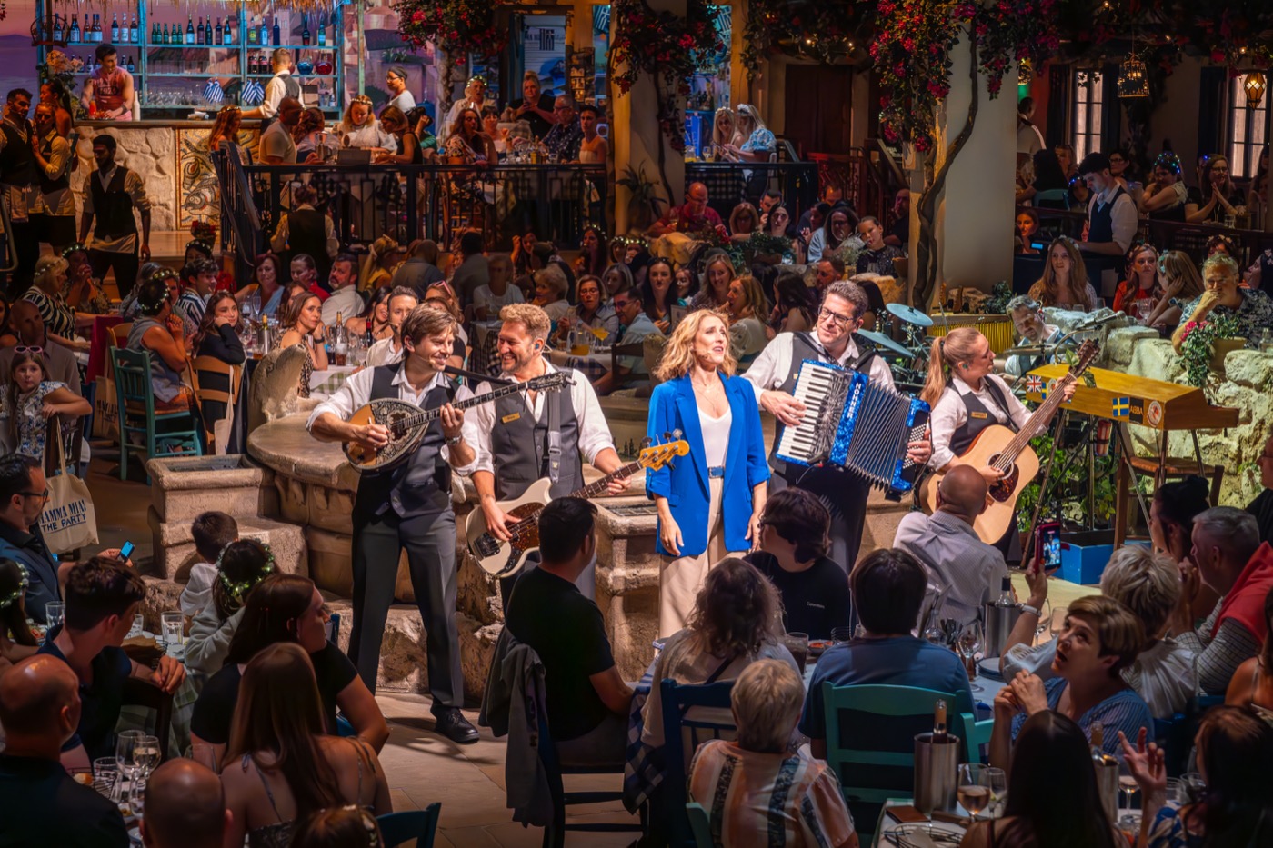 A lively performance at Mamma Mia! The Party with musicians and actors entertaining a vibrant crowd in a festive Greek setting.