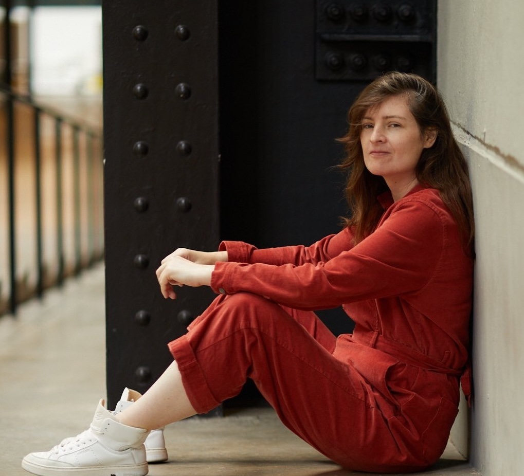 Madeleine Brettingham sitting casually in a red jumpsuit and white sneakers, exuding creative energy for Made in Shoreditch feature