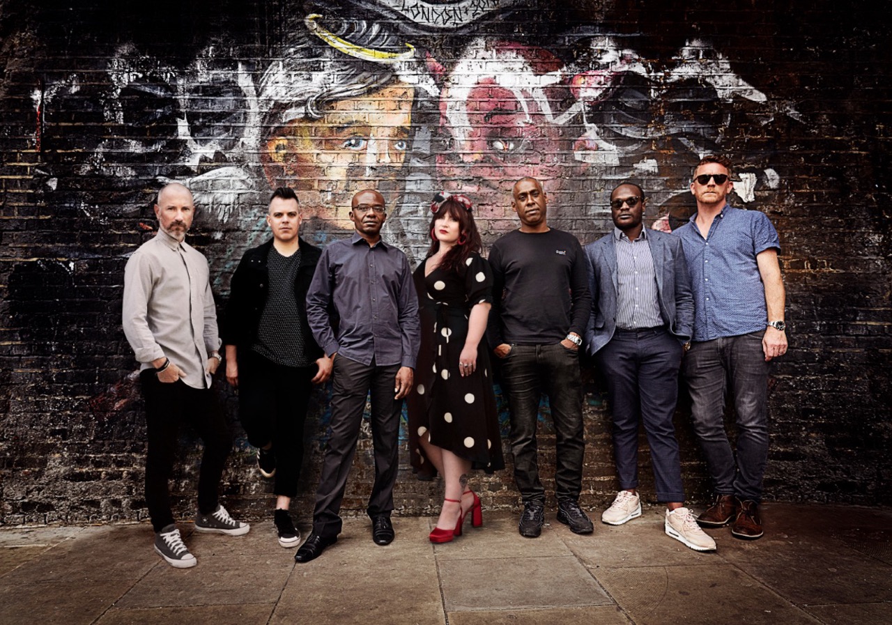 The Amy Winehouse Band poses against a graffiti-covered London wall, celebrating their legacy and upcoming tribute performance.