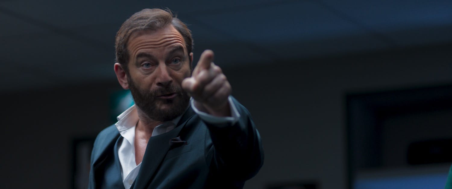 Jason Isaacs mid-scene, framed by shadow and fluorescent spill